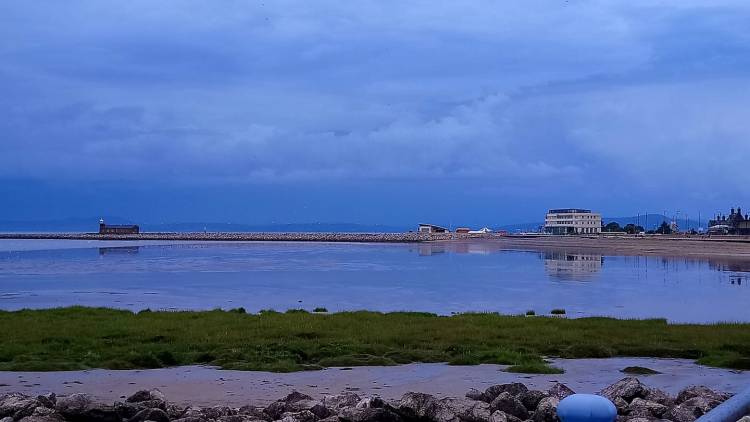 Striking peaceful and scenic image of morecambe bay and the midland hotel. Reflections of the hotel and stone jetty cafe in the calm glass like water. Sky as after a storm, clouds dissipating and deep blue sky.